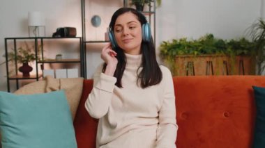 Happy overjoyed brunette woman in wireless headphones relaxing sitting on couch at home apartment choosing listening favorite energetic disco dancing music. People weekend daytime leisure activities