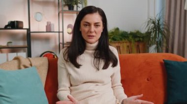 What. Why. Young woman raising hands in indignant expression asking reason of failure demonstrating disbelief irritation by troubles at home apartment. Brunette girl in living room sitting on sofa