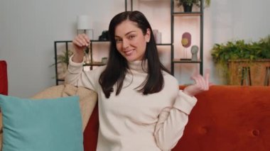 Young woman real estate agent lifting hand showing the keys of new home house apartment, buying or renting property, mortgage loan at modern home apartment indoors. Adult girl in room sitting on sofa