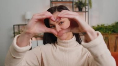 I love you. Happy brunette woman at home living room couch makes symbol of love, showing heart sign to camera, express romantic feelings express sincere positive feelings. Charity, gratitude, donation