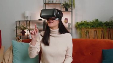 Young woman using virtual reality futuristic technology VR app headset helmet to play simulation 3D 360 video game, watching film movie at modern home apartment. Blonde girl in goggles sitting on sofa