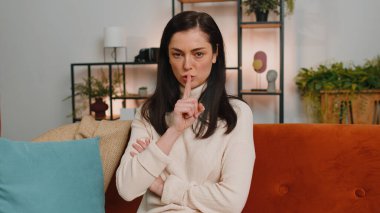 Shh be quiet, hush please. Young woman with brunette hair presses index finger to lips makes silence hush gesture sign do not tells gossip secret. Girl at modern home living room sitting on couch