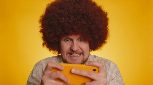 Worried bearded man with afro hairstyle coiffure enthusiastically play racing or shooter video games on smartphone. Hipster guy use mobile phone gadget app with drive simulator on yellow background