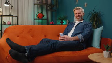 Mature senior bearded man lying on sofa uses mobile phone smiles at night evening home. Mature guy texting share messages content on smartphone social media applications online watching relax movie