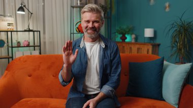 Orta yaşlı bir adam kameraya dostça gülümsüyor ve el sallayarak merhaba, selam veriyor ya da hoşça kal diyor, misafirperver bir ifadeyle karşılıyor. Olgun, yaşlı, gri saçlı adam evde, kanepede yatıyor.