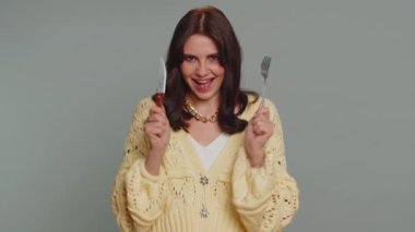 Ready to eat. Hungry young woman in cardigan waiting for serving dinner dishes with cutlery, will appreciate delicious restaurant meal menu. Girl holding fork and knife on gray studio background