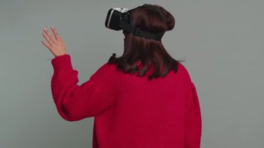 Woman using headset helmet to play simulation game app online. Watching virtual reality 3D 360 video content. Girl in VR goggles isolated on studio gray background. Futuristic technology. Rear view