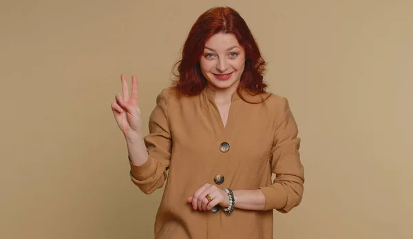 Hipster redhead woman in blouse showing victory v sign, hoping for success and win, doing peace gesture, smiling with kind optimistic expression. Young adult girl isolated on beige studio background