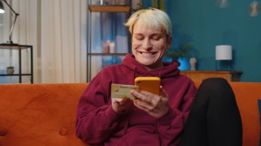 Kısa saçlı genç bir kadın kredi kartı ve akıllı telefon kullanarak para transfer ediyor, online alışveriş yapıyor, ev içinde yemek siparişi veriyor. Kız kanepede yatıyor.