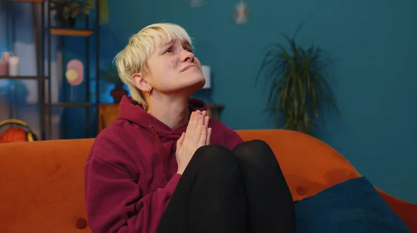 Please, God help. Woman with short blonde hair praying sincerely with folded arms, looking upward and making wish, asking with hopeful imploring expression, begging apology. girl at home living room