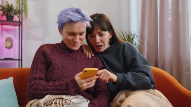 两个已婚女同性恋夫妇或朋友坐在家里客厅的沙发上 使用智能手机 快乐的女孩家庭得到了积极的测试结果 庆祝胜利 拥抱彼此 — 图库视频影像