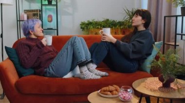 Two young smiling lesbian women family couple or girls friends drinking coffee or tea and talking at home morning living room, lying on sofa. LGBT same-sex people. Enjoying happy breakfast together