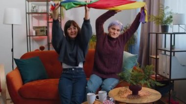 Two lesbian women family couple or girl friends holding LGBT people gay pride flag in hands at home living room. Gay, bisexual, transgender social movements. Concept of happiness freedom love same-sex