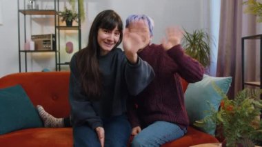Two lesbian women family couple or girls friends smiling friendly at camera. Girls waving hands gesturing hello, hi, greeting or goodbye, welcoming with hospitable expression at home room. LGBT people