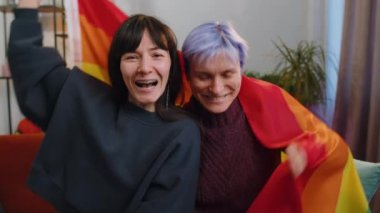 Homosexual lesbian couple sitting at home room couch next to each other, holding LGBT flag. Smiling girls friends. Pregnant woman from IVF. Lesbian family. Concept of happiness freedom love same-sex