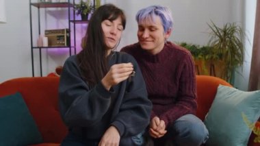 Two young lesbian women family couple or girls friends lifting hand showing the keys of new home house apartment, buying or renting real estate property, mortgage loan at living room. LGBT people