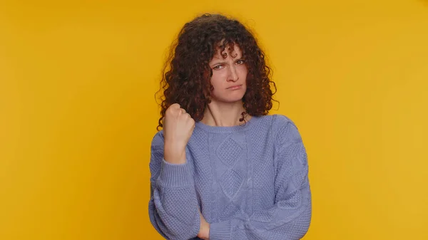 Aggressive angry curly haired woman trying to fight at camera, shaking fist, boxing with expression, punishment, disappointment, rage, quarrel. Young teen girl isolated on yellow studio background