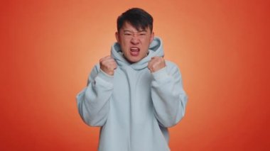 Asian man in hoodie screams yell from stress tension problems feels horror hopelessness fear panic surprise shock expresses gestures rage. Handsome chinese guy isolated on orange studio background
