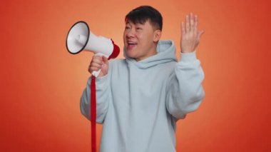 Asian man talking with megaphone, proclaiming news, loudly announcing advertisement, warning using loudspeaker to shout speech, pointing empty place. Copy-space. Handsome guy on orange wall background