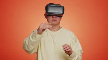 Asian man using headset helmet to play simulation game app online. Watching virtual reality 3D 360 video content. One guy in VR goggles isolated on studio orange background. Futuristic technology