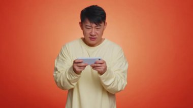 Worried funny addicted asian man enthusiastically playing racing video games on mobile phone. Handsome adult guy using smartphone gadget app with drive simulator isolated on orange studio background