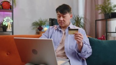 Asyalı adam para transfer ederken kredi kartı ve dizüstü bilgisayar kullanıyor, online alışveriş yapıyor, eve yemek siparişi veriyor. Oturma odasında oturan yetişkin bir adam. İçerideki koltukta oturuyor.