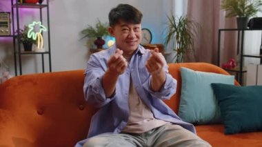 Cheerful happy rich asian man showing wasting, throwing money around hand gesture, more tips earnings, big profit, win lottery, body language, share, celebrate. Guy at home in living room apartment