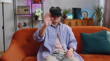 Asian man using virtual reality futuristic technology VR app headset helmet to play simulation 3D 360 video game, watching film movie at modern home apartment. Blonde guy in goggles sitting on sofa