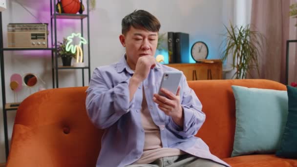 Asiatisk Mann Som Sitter Sofaen Smiler Mobilen Hjemme Kinesisk Voksen – stockvideo