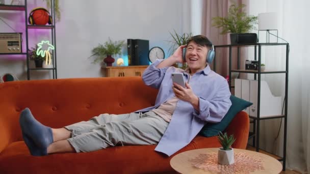 Happy Overjoyed Chinese Asian Man Wireless Headphones Relaxing Sits Couch — 图库视频影像