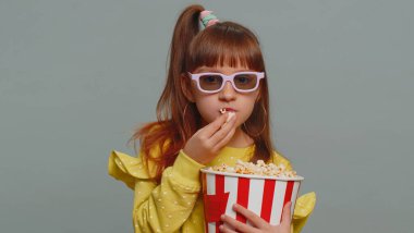 Excited young toddler school girl in 3D glasses eating popcorn, watching interesting tv serial, sport game, film, online social media movie content. Teen female child kid on studio gray background