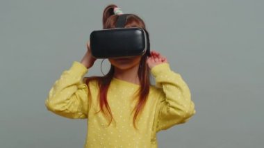 Sanal gerçeklik gözlüklü küçük kız, simülasyon oyunu oynamak için kulaklık uygulaması kullanıyor. Sanal gerçeklik 3D 360 videosu izliyoruz. Stüdyonun gri arka planında izole edilmiş genç kız çocuk. Küçük çocuklar