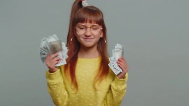 Rich pleased young toddler school girl in glasses waving money dollar cash banknotes bills like a fan, success, lottery winner, big income, wealth. Teen female child kid on studio gray background