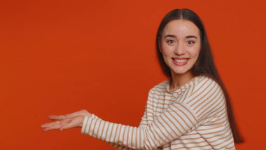 Lovely millennial pretty woman showing thumbs up and pointing empty place, advertising area for commercial text, copy space for goods promotion. Young girl. Studio shot indoors on red wall background
