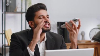 Indian businessman working, having mobile phone loudspeaker talk at home office desk with laptop. Bearded professional freelance man holding smartphone using messenger chat apps. Employment occupation