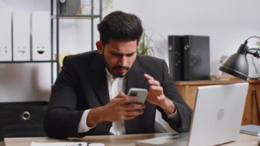 Indian businessman in suit use smartphone typing browsing, loses becoming surprised sudden lottery results, bad news, fortune loss fail at home office desk workplace. Bearded freelancer worker man