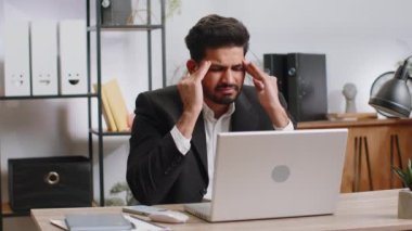 Tired ill indian businessman suffering from headache problem tension and migraine, stress at home office desk workplace. Bearded freelancer broker worker man works on laptop notebook. Overworking