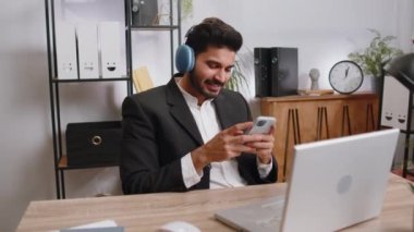 Happy relaxed overjoyed indian businessman working on laptop computer at home office wearing headphones listening favorite energetic disco music on smartphone. Freelancer man relaxing, taking a break