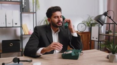 Bearded indian businessman working, having talk with client on retro old-fashioned wired telephone at home office workplace desk. Salesman in formal suit. Remote distant job. Employment occupation