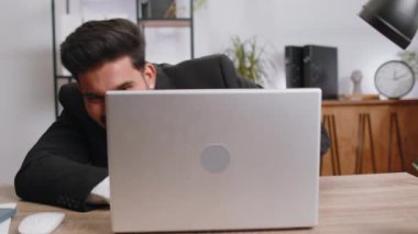 Indian businessman hiding behind laptop computer, making funny face, fooling around, putting his tongue out remote working disrespecting someone at office desk workplace. Professional freelancer man