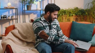 Indian man freelancer opens laptop, starts working education on notebook, sends messages, makes online purchases, watching movies at home living room apartment. Hindu guy sitting on couch. Lifestyles