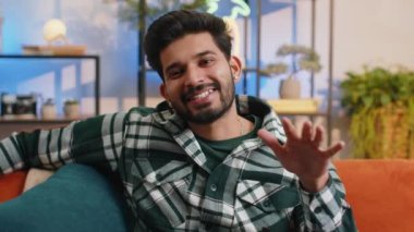 POV of indian man looking at camera, making video webcam conference call with friends or family, enjoying pleasant conversation at home room apartment. Hindu guy sits on sofa laughing, waving hello