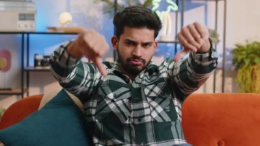 Dislike. Upset indian man showing thumbs down sign gesture, expressing discontent, disapproval dissatisfied bad work at modern home apartment indoors. Displeased hindu guy in living room on sofa