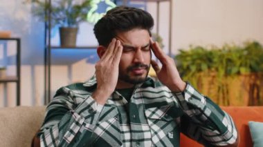 Displeased indian man rubbing temples to cure headache problem, suffering from tension and migraine, stress grimacing in pain high blood pressure at home room apartment. Hindu guy on couch. Lifestyles