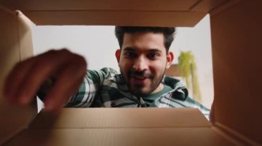 Inside view from box. Indian man unpacking delivery parcel at home. Smiling satisfied hindu guy shopper, online shop customer opening cardboard receiving purchase gift by fast postal shipping order