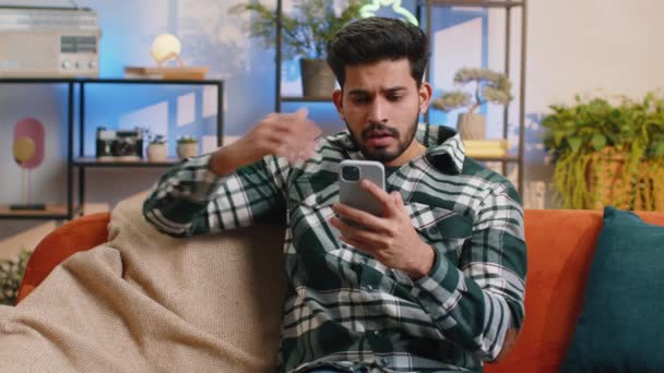 悲しい不満インドの男は スマートフォンの入力ブラウジングを使用して 突然宝くじの結果 悪いニュース 幸運損失 ウイルスに驚いて失う 若いですHindu男でホームリビングルームオンソファ — ストック動画