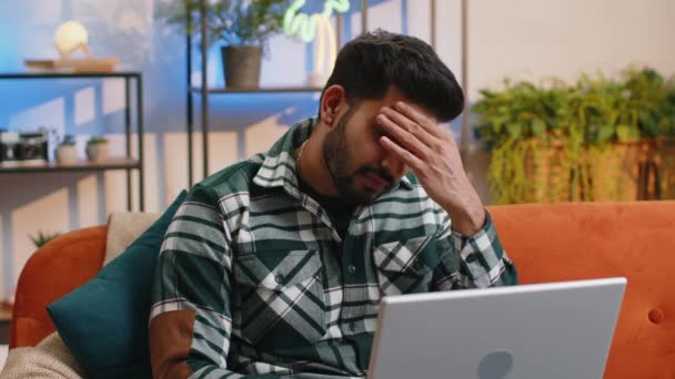 疲れたフリーランスのインド人男性は 自宅の部屋で頭痛の問題の緊張と片頭痛 ストレスに苦しんで ラップトップを使用します ヒンズー教の男はノートブックで働き メッセージを送り オンラインで購入し 映画を見ます — ストック動画