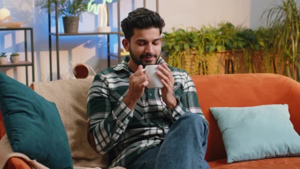 家庭のリビングルームのアパートで暖かいコーヒーやハーブティーを飲んでハンサムな笑顔のインド人男性 休憩時間だ ヒンドゥ教徒の男は快適なリラックスした朝にソファに座って楽しむ ライフスタイル — ストック動画