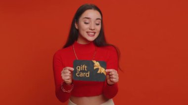 Young woman in crop top showing pointing on gift discount certificate voucher coupon for store holidays sale. Lovely girl with shopping christmas surprise card isolated alone on red studio background