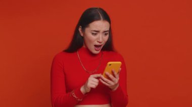 Pretty woman playing on smartphone typing browsing, loses becoming surprised sudden lottery results, bad fortune, loss, game fail, bankruptcy. Young girl isolated alone on red studio wall background